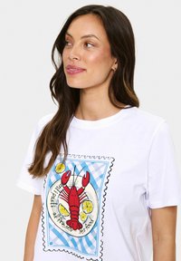 T-shirt en coton blanc arborant un graphique de homard en rouge, entouré de citrons jaunes et d'une bordure à carreaux bleus. Manches courtes, col rond.