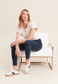 Wit T-shirt met korte mouwen, marineblauwe broek en witte sneakers met zwarte accenten. Model zit op een wit stoeltje met gouden accenten tegen een beige achtergrond.