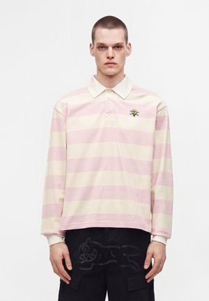 Giovane uomo che indossa una polo a righe rosa e crema a manica lunga con un piccolo logo floreale ricamato e pantaloni neri con design a cartone animato.
