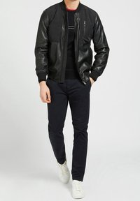 Giubbotto bomber in pelle nera con zip frontale, polsini a coste e taschino sul petto. Indossato sopra una maglietta nera a maniche lunghe e abbinato a pantaloni scuri e sneakers bianche.