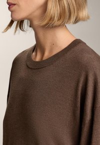 Barna kötött pulóver, amely puha tapintású, kerek nyakkivágással és laza fazonnal rendelkezik, finom fénnyel, sima felületet és kényelmes viseletet biztosít.