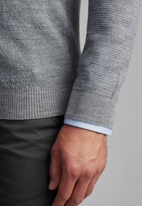 Pull en tricot gris avec un tissu texturé et des poignets côtelés, superposé sur une chemise à col bleu clair, partiellement visible au niveau des manches.
