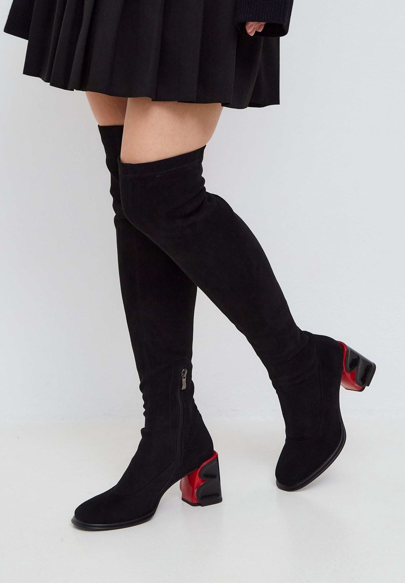 Botas negras de ante por encima de la rodilla con punta redondeada y un tacón grueso negro con un acento rojo. Cierre de cremallera lateral para facilitar su uso.