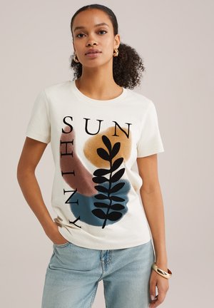 T-shirt en coton de couleur crème avec du texte noir en gras "SUN SHINY" et des formes colorées abstraites en beige, bleu et rouille avec un motif de feuille.