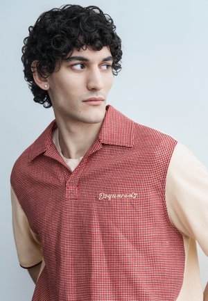 Jeune homme aux cheveux bouclés portant un polo à motif pied-de-poule rouge et beige, regardant vers sa gauche sur un fond uni.