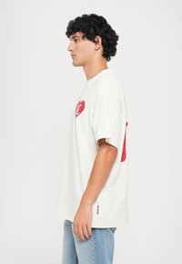 Off-white cotton t-shirt with short sleeves, featuring a red heart graphic and text on the front. Model wears blue jeans, standing sideways.