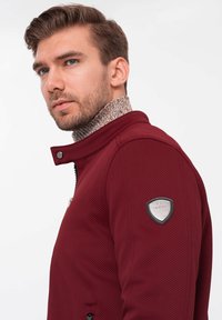 Burgundy zip-up jacka med texturerad tyg, hög krage och kontrasterande stickad underlayer. Har en silverfärgad logotyp på vänster arm.