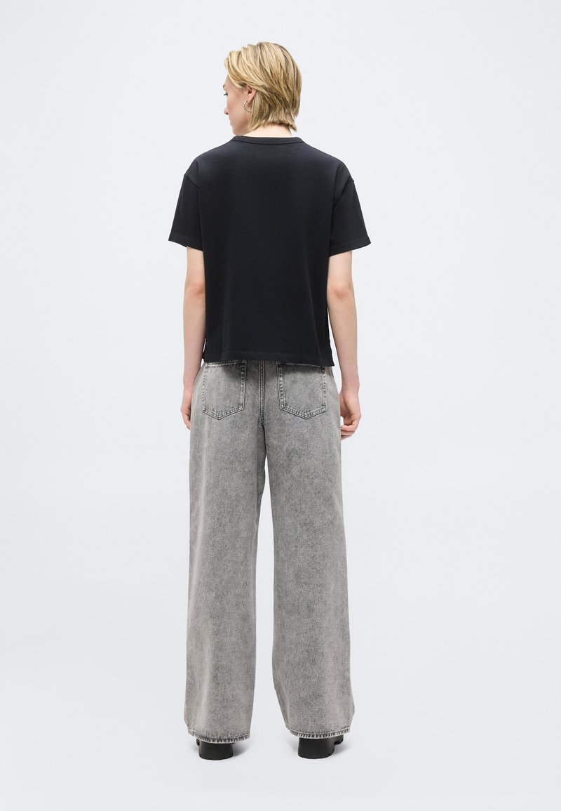 Personne aux cheveux blonds courts portant un t-shirt noir à manches courtes et un jean gris à jambes larges, debout de dos sur un fond blanc uni.