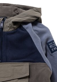 Polarowy dres z połączeniem tkanin w kolorze granatowym, szarym i beżowym. Posiada zamek błyskawiczny, kaptur oraz przednią kieszeń z klapą z detalem logo.