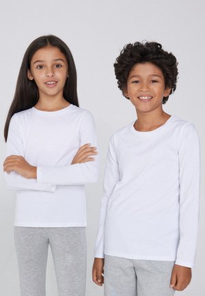 Zwei Kinder tragen langärmelige weiße Hemden. Das Mädchen hat glattes Haar und graue Hosen, während der Junge lockige Haare und graue Hosen hat.