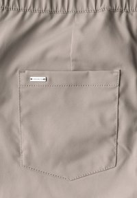 Beige Shorts mit elastischem Bund, ausgestattet mit einer Gesäßtasche. Auf der Tasche befindet sich ein silbernes Etikett, das angenäht ist. Glatte Textur, lässiges Design.