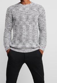 Mann in einem grau-weißen Strickpullover und schwarzen Hosen, mit den Händen in den Taschen, vor einem einfarbigen hellgrauen Hintergrund stehend.