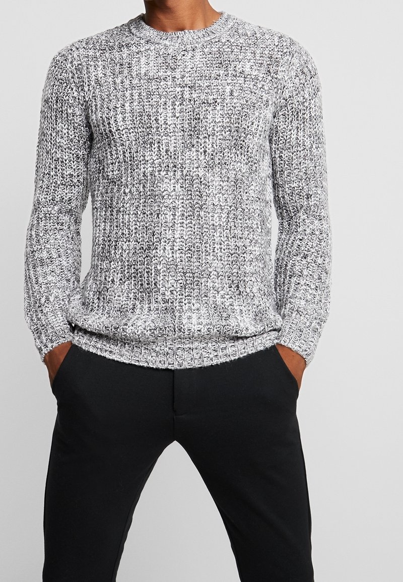 Mann in einem grau-weißen Strickpullover und schwarzen Hosen, mit den Händen in den Taschen, vor einem einfarbigen hellgrauen Hintergrund stehend.