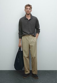 Dunkelgrauer Kragensweater, hellkhakifarbene, gefaltete Hose und schwarze Tragetasche. Das Modell trägt dunkle Schuhe mit einer strukturierten Sohle auf einer dunklen Oberfläche.