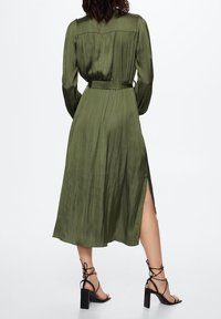 Robe verte olive à manches longues avec une taille cintrée, des détails plissés au dos et une fente sur le côté ; portée avec des sandales à talons hauts noires à lanières.