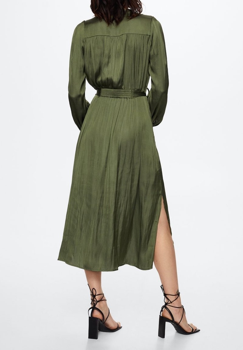Robe verte olive à manches longues avec une taille cintrée, des détails plissés au dos et une fente sur le côté ; portée avec des sandales à talons hauts noires à lanières.