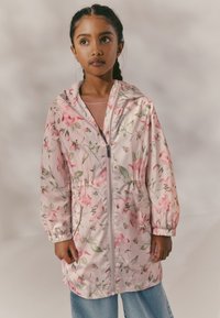 Veste de pluie légère rose à imprimé floral, fermeture éclair, capuche et poignets élastiques. Associée à un jean bleu clair. Texture lisse.