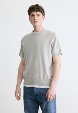 Joven con cabello rizado vistiendo un suéter de manga corta gris claro sobre una camiseta blanca y jeans azules, de pie frente a un fondo liso.