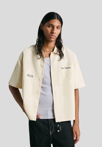 Una giovane persona con capelli lunghi e scuri indossa una camicia a maniche corte di colore crema sopra una canotta grigia e jeans scuri con accessori a catena.