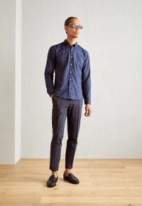 Navy button-up shirt, slim-fit gray trousers, black loafers. Sleek sunglasses. Smooth fabric, tailored design, and modern silhouette.