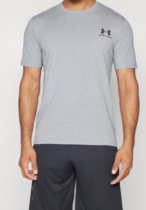 Mann trägt ein hellgraues Under Armour Kurzarm-T-Shirt und schwarze Sportshorts, steht mit locker hängenden Armen an den Seiten.