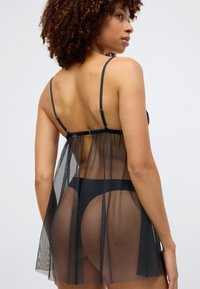 Svart gjennomsiktig mesh-lingerie med justerbare stropper, med en tettsittende bodice og flytende bakpaneler som draperer til midt på låret.