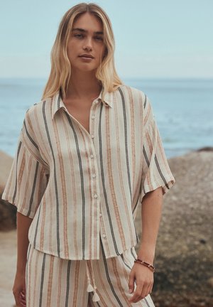 Femme debout au bord de la mer portant une chemise beige à rayures manches courtes boutonnée et un pantalon assorti avec un cordon à pompons, bracelet marron au poignet.