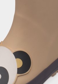 Κοντινό πλάνο ενός μπεζ παιδικού παπουτσιού με λεπτομέρεια προσώπου και αυτιών πάντα σε μαύρο και άσπρο στο μπροστινό μέρος.