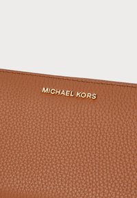 Rjavo teksturirano usnjeno torbico z vtisnjenim zlatim logotipom "MICHAEL KORS" na sprednji strani, ki prikazuje eleganten, minimalističen dizajn.