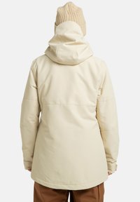Osoba w kremowej, zimowej kurtce z kapturem i beżowej czapce dzianinowej, przedstawiona od tyłu na jednolitym tle.