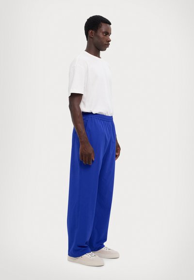 T-shirt blanc à manches courtes, pantalon bleu ample, taille élastique, tissu texturé et baskets de couleur claire sur un fond neutre.