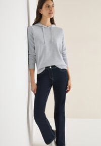 Felpa grigia con cappuccio, maniche lunghe e polsini a coste; abbinata a jeans scuri a zampa con cuciture a contrasto e sneakers bianche.