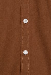 Camicia in tessuto marrone con texture, caratterizzata da una fila di bottoni bianchi lungo il pattina, con dettagli di cucitura visibili.