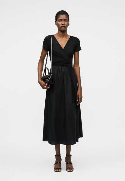 Žena v čiernych zavíjacích šatách, s čiernou kabelkou na ramene a čiernymi pásikovými lodičkami, stojaca pred jednoduchým bielym pozadím.