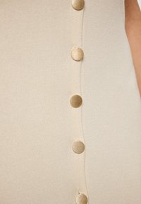 Il tessuto a maglia beige presenta una fila verticale di cinque bottoni dorati, mettendo in risalto una texture liscia e un leggero luccichio.