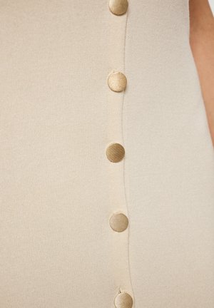 Il tessuto a maglia beige presenta una fila verticale di cinque bottoni dorati, mettendo in risalto una texture liscia e un leggero luccichio.