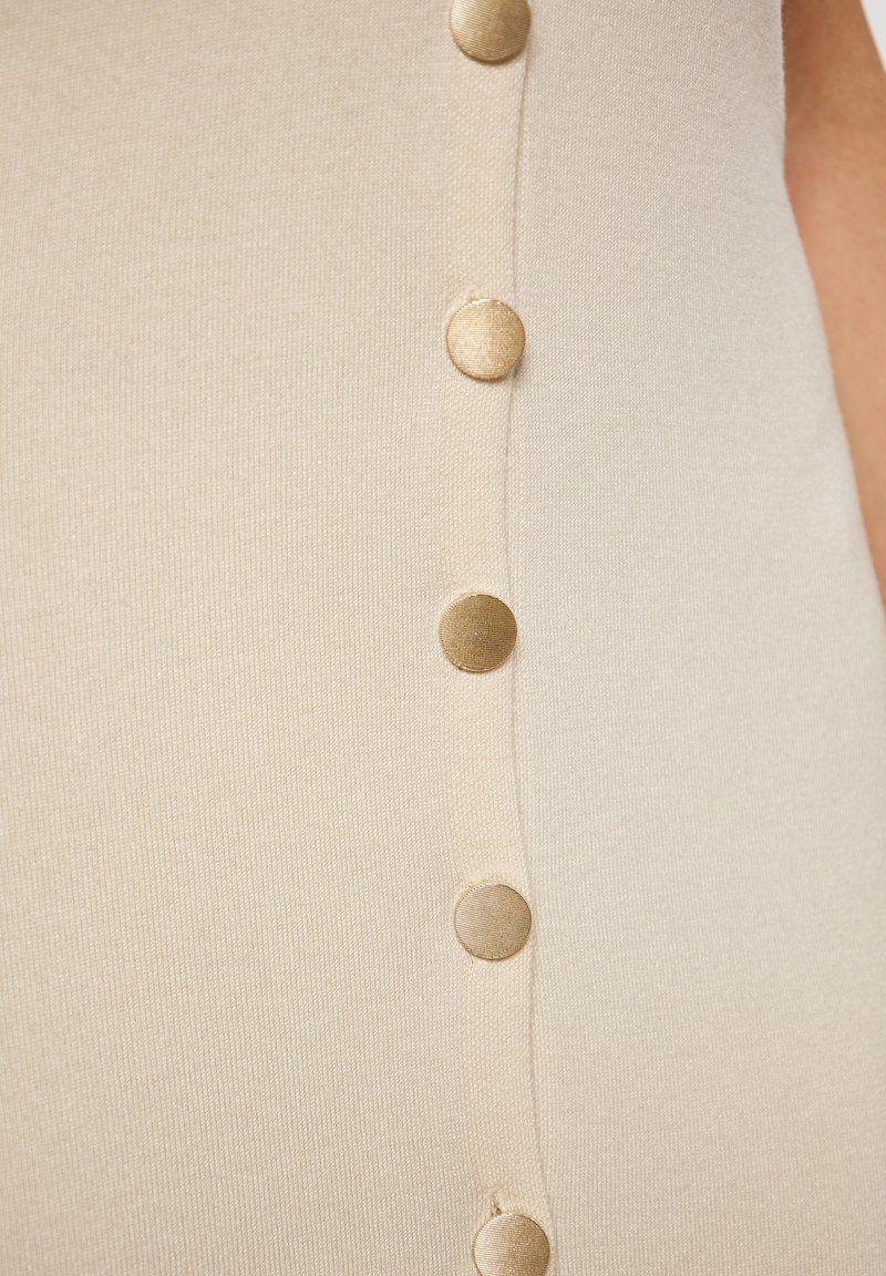 Il tessuto a maglia beige presenta una fila verticale di cinque bottoni dorati, mettendo in risalto una texture liscia e un leggero luccichio.