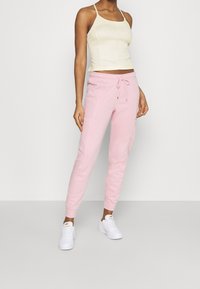 Light pink jogger pants with elastic waist, drawstring detail, and side pockets. Worn with a fitted cream tank top and white sneakers.