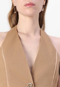Halter-necken yläosa, vaalea ruskea, syvällä V-kaula-aukolla, ohut beige reunus sekä etupuolella nappiyksityiskohta, jossa pyöreä koriste.