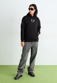 Sort hættetrøje med BMW Motorsport-logo, grå denimbukser og sorte sporty sko. Modellen har store solbriller på.