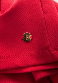 Červená látka s hladkým povrchom, s malým zlatým okrúhlym logom s vystúpeným dizajnom „B“. Viditeľné detaily šitia na okrajoch.