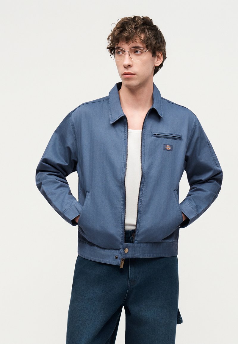 Jonge man met bril die een blauwe jas met rits draagt over een wit overhemd en donkere spijkerbroek, staand met handen in de zakken.