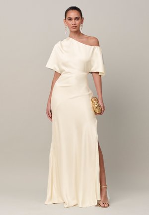 Mujer con un vestido de satén crema de un solo hombro, sosteniendo un bolso de mano dorado, con pendientes dorados y tacones abiertos sobre un fondo liso beige.