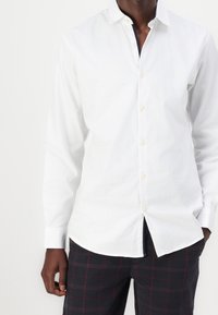 Camisa blanca de botones con mangas largas, textura suave, cuello estándar y botones beige, llevada con pantalones a cuadros oscuros con acentos rojos.