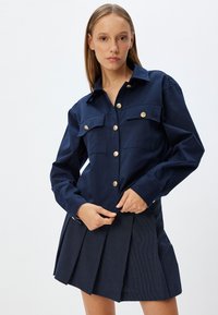 Camicia button-up blu navy con due tasche frontali e bottoni oro, abbinata a una gonna a pieghe a righe.