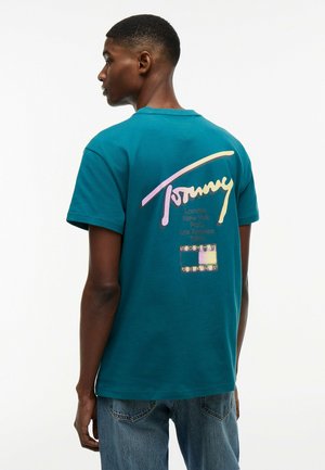 Joven de espaldas vistiendo una camiseta azul verdoso de Tommy Jeans con logo en colores pastel y nombres de ciudades impresos en la espalda, combinado con vaqueros azules.