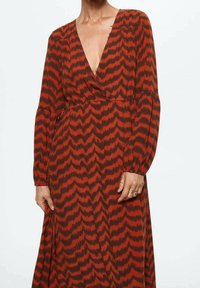Frau trägt ein langärmliges Kleid mit tiefem V-Ausschnitt und einem rot-schwarzen Zickzackmuster, das einen Teil ihres Halses, ihrer Brust und ihrer Hände zeigt.