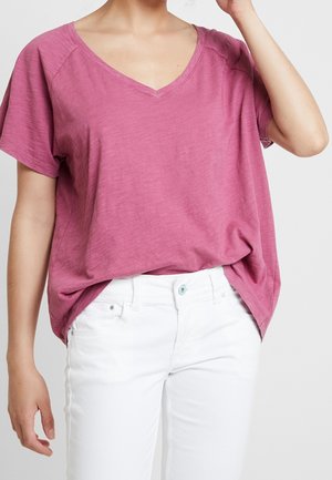 Person wearing a loose pink V-neck short-sleeve top tucked slightly into high-waisted white pants against a light background.
