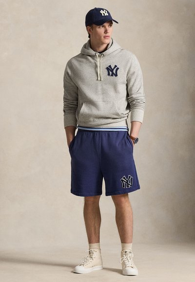 Γκρι φούτερ με κουκούλα και ναυτικό λογότυπο, συνδυασμένο με ναυτικό σορτς με τυπωμένο λογότυπο και μπεζ αθλητικά παπούτσια υψηλού τύπου. Καθημερινό αθλητικό σύνολο.