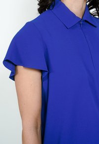 Blå top med krave og korte, flæsekantede ærmer. Stoffet virker glat og let. Lynlåsdetalje synlig ved halsen.